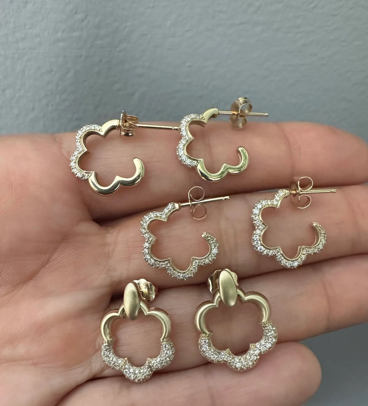 Onirikka Gold flower-shaped yellow gold earrings on a hand against a gray background