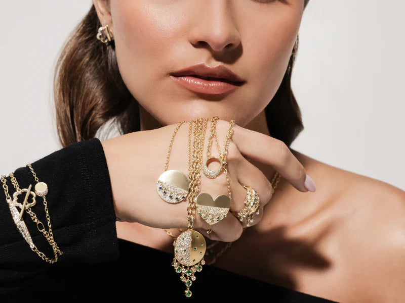 Close-up of a woman's hand wearing showing Onirikka Heart Hemline Medallion in satin 14k yellow gold with diamonds and emeralds.