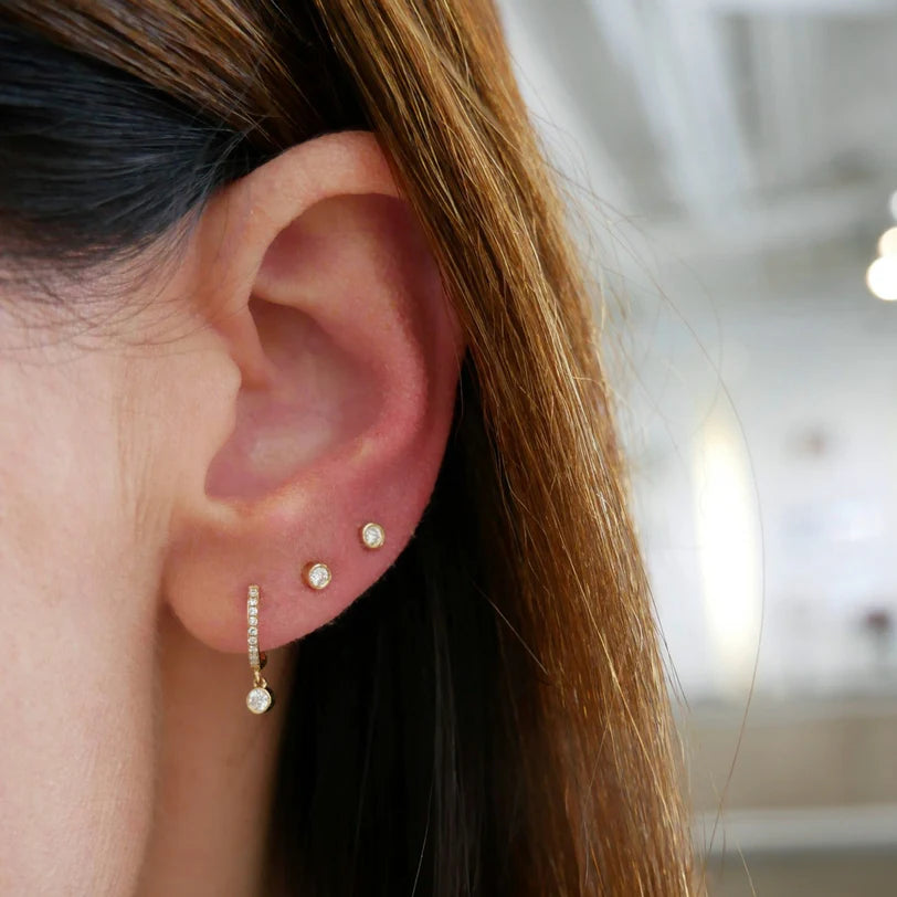 EF Collection Diamond Bezel Drop Mini Huggie Earring in yellow gold styled on models ear with stud earrings. 