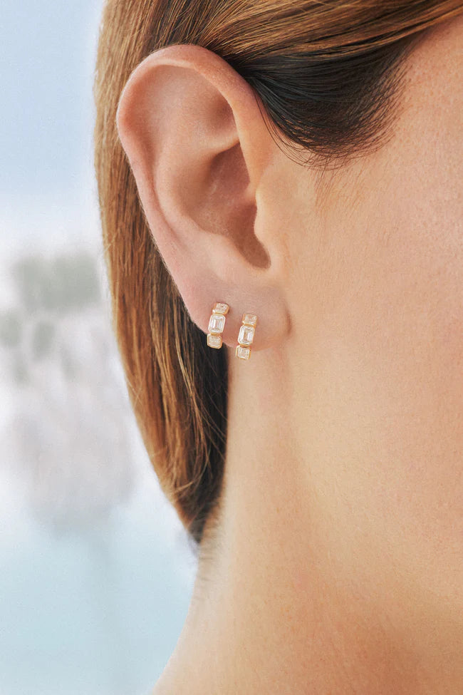 Emerald Cut Diamond Bezel Mini Huggie Earrings in yellow gold on models ear. 